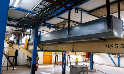 Interior de nave industrial con cinta transportadora elevada, conductos metálicos y estructura azul de soporte.