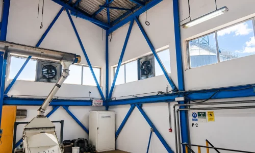 Interior de nave industrial con estructura metálica azul, maquinaria de cribado y extracción, ventanas altas y sistema de ventilación.