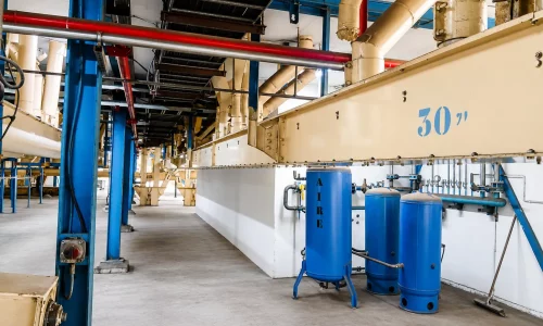 Interior de nave industrial con conductos metálicos, estructura azul y depósitos de aire comprimido junto a una línea de maquinaria.